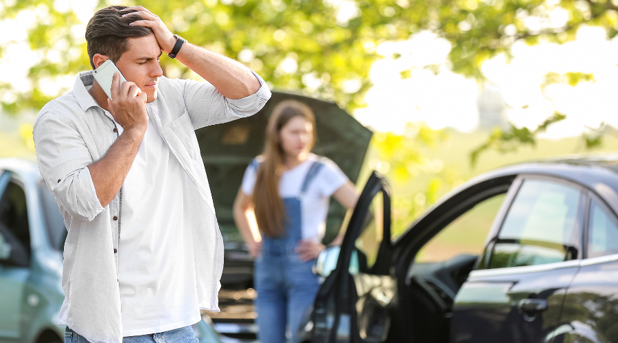 Man on phone at the scene of a car accident with woman in background with hands on hips.