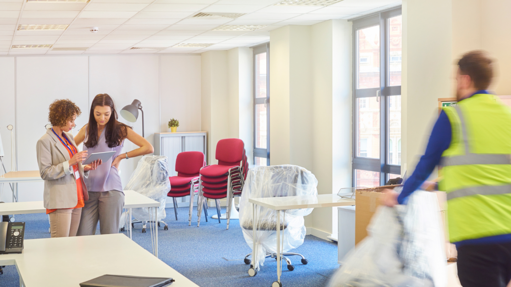 two woman looking a a clipboard, standing in a room with office furniture.