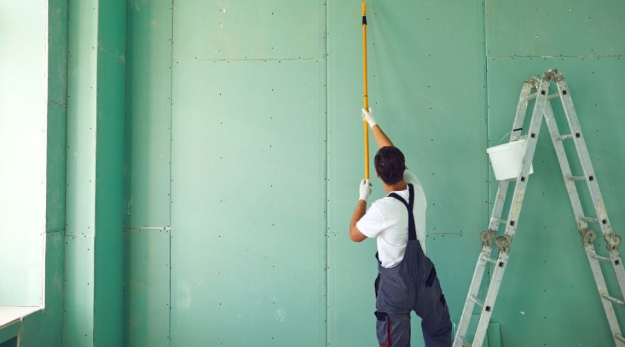 A painter in overalls uses a paint roller to paint a ceiling green.