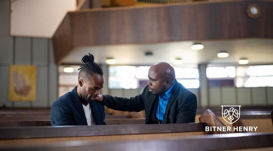 A pastor leads his congregation in safety by comforting a congregant. The Bitner Henry logo is in the bottom right.
