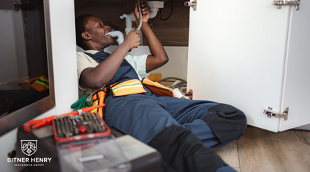 A plumber fixes the pipes below a sink with the bitner henry logo in the bottom left.