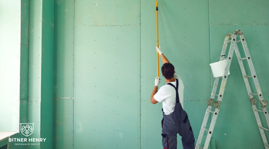A painter in overalls uses a paint roller to paint a ceiling green.